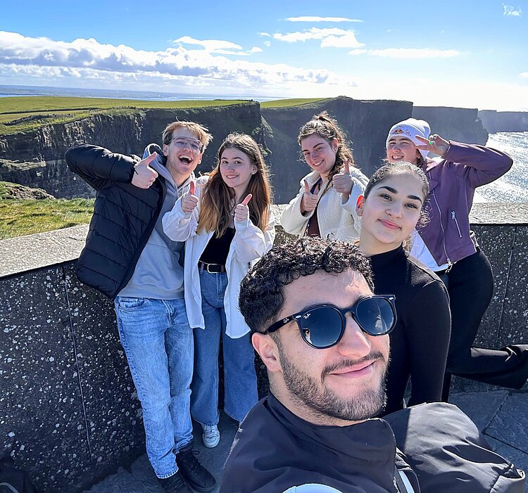 „Man muss reisen, um zu lernen“: Auslandspraktikum mit dem Förderprogramm Erasmus+ Sechs junge Menschen stehen lächelnd und mit Daumen hoch an den Cliffs of Moher in Irland. Im Hintergrund sind die steilen Küstenfelsen und das Meer bei strahlend blauem Himmel zu sehen. Eine Person im Vordergrund macht ein Selfie der Gruppe.