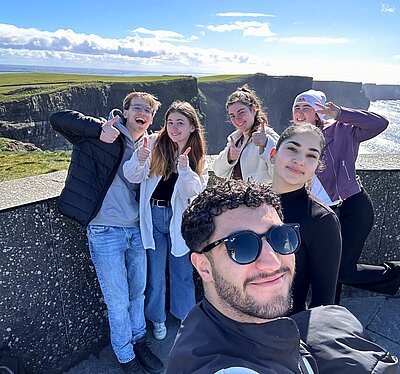Sechs junge Menschen stehen lächelnd und mit Daumen hoch an den Cliffs of Moher in Irland. Im Hintergrund sind die steilen Küstenfelsen und das Meer bei strahlend blauem Himmel zu sehen. Eine Person im Vordergrund macht ein Selfie der Gruppe.
