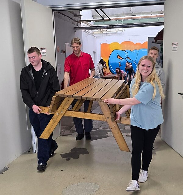 Die Schülerinnen und Schüler der Euro Akademie tragen einen großen hölzernen Tisch mit integrierter Sitzbank.