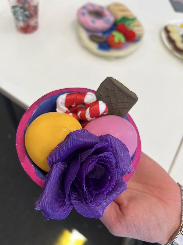 Eine Hand hält ein rundes, buntes Objekt, das wie eine kleine Schale gestaltet ist. Darin befinden sich eine künstliche lila Blume, zwei runde Kugeln in Gelb und Rosa, ein rechteckiges Stück in Schokoladenoptik sowie ein rot-weiß gestreiftes Element, das an eine Zuckerstange erinnert. Im Hintergrund sind auf einem weißen Tisch weitere bunte, modellierte Objekte unscharf zu erkennen.