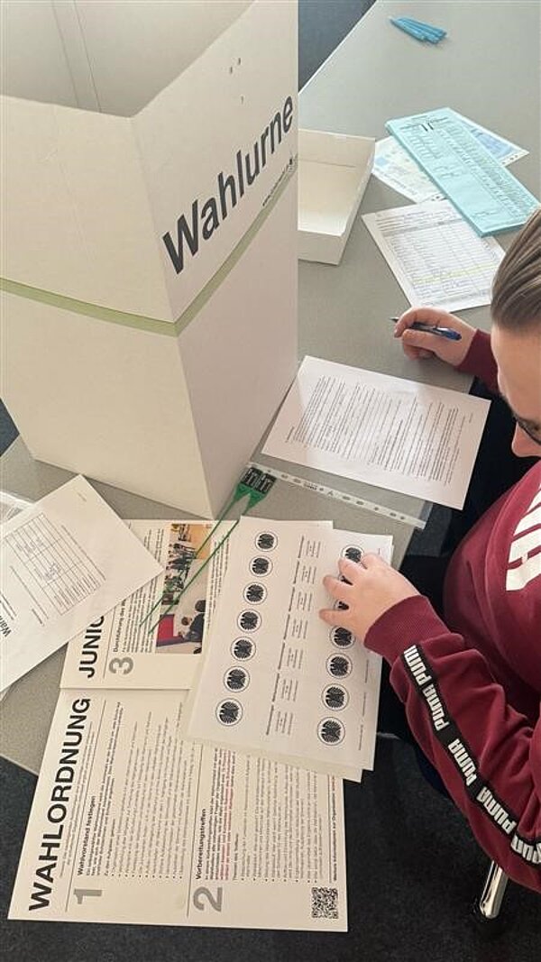 Testwahlen an der Euro Akademie Aschaffenburg am 19. Februar 2025 Eine Nahaufnahme eines Tisches mit Wahlunterlagen, darunter ein Stimmzettel mit offiziellen Siegeln und die Wahlordnung. Daneben steht eine Wahlurne.