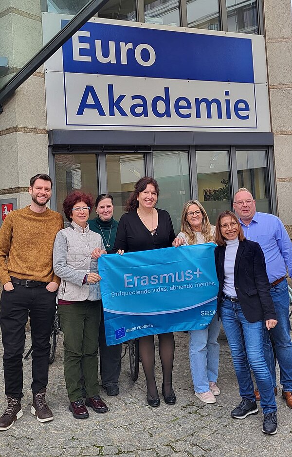 Eine Gruppe von sieben Personen steht vor der Euro Akademie und hält eine Erasmus+ Flagge. Die Gruppe besteht aus Männern und Frauen unterschiedlichen Alters, die lächelnd in die Kamera schauen. Im Hintergrund ist das Schild der Euro Akademie sichtbar.