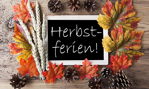 Ein Schiefertafel-Schild mit der Aufschrift ‚Herbstferien!‘, umgeben von bunten Herbstblättern, Tannenzapfen und einer Ährenrispe.