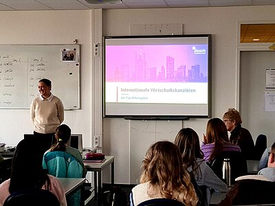 Eine Person steht lächelnd vor einer Klasse und präsentiert eine Folie mit dem Titel „Internationale Wirtschaftskanzleien als Top-Arbeitgeber“ von Desch Partners. Im Raum sitzen mehrere Schülerinnen an Computertischen.