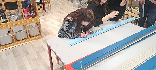 Eine junge Frau macht mit einem Stift Markierungen auf einer hellblauen Holzlatte.