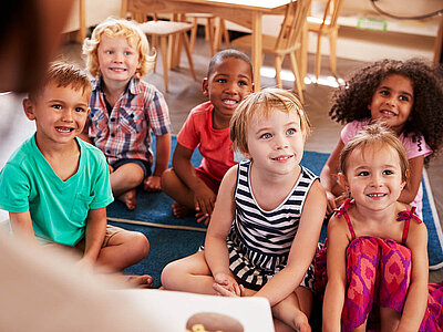 Sechs Kinder sitzen auf einem Teppich in einem Klassenzimmer und schauen aufmerksam nach vorne. Die Kinder wirken fröhlich und interessiert.
