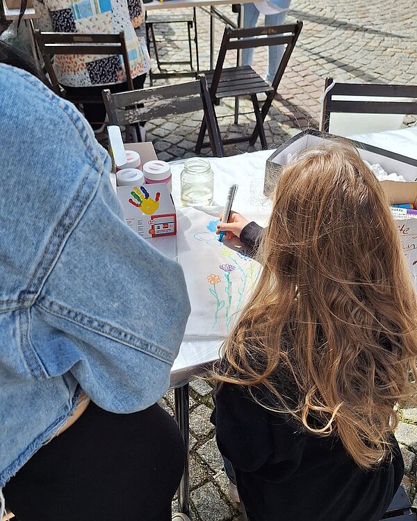 Ein Kind malt mit bunten Stiften Blumen auf ein Blatt Papier bei der Bastelstation der Euro Akademie. Daneben stehen Gläser mit Wasserfarben und weitere Bastelmaterialien. Die Aktion findet im Rahmen des Frühlingsfests statt.