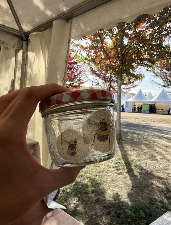 Eine Hand hält ein kleines Einmachglas mit zwei Bildern von Bienen. Im Hintergrund sind weiße Zelte und Bäume zu sehen.