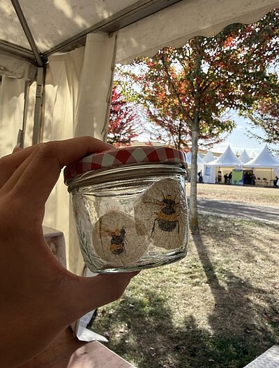 Eine Hand hält ein kleines Einmachglas mit zwei Bildern von Bienen. Im Hintergrund sind weiße Zelte und Bäume zu sehen.