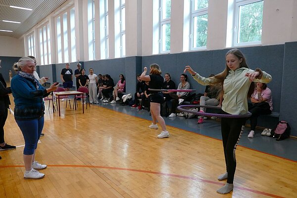 In einer Turnhalle üben zwei Personen das Hula-Hoop-Tanzen. Eine ältere Frau in blauem Outfit steht links und schaut auf ein Smartphone, während eine jüngere Frau in einem hellen Hoodie und schwarzer Leggins den Hula-Hoop-Reifen um ihre Taille kreisen lässt. Im Hintergrund sitzen weitere Personen auf Bänken und beobachten.