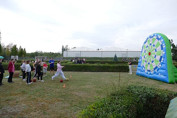 Eine Person ist kurz davor, einen Ball auf ein großes, aufblasbares Dartbrett im Freien zu kicken. Im Hintergrund sind weitere Personen zu sehen, die der Aktivität zuschauen oder sich unterhalten. Das Dartbrett ist blau-weiß mit Zahlen markiert.