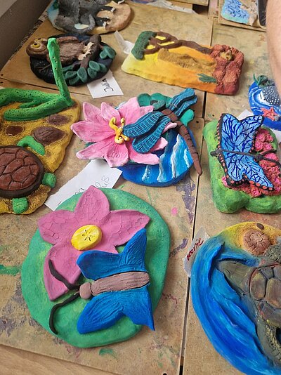 Eine Sammlung von mehreren farbenfrohen Tonreliefs auf einer Holzplatte. Die Motive umfassen eine Schildkröte, Blumen mit Schmetterlingen oder einer Libelle, einen Vogel, eine Eule und vieles mehr.