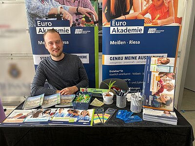 An einem Messestand sitzt ein lächelnder Mann. Hinter ihm stehen Roll-ups der Euro Akademien Meißen und Roesa. Auf dem Tisch vor ihm liegen Werbematerialien der Euro Akademie.