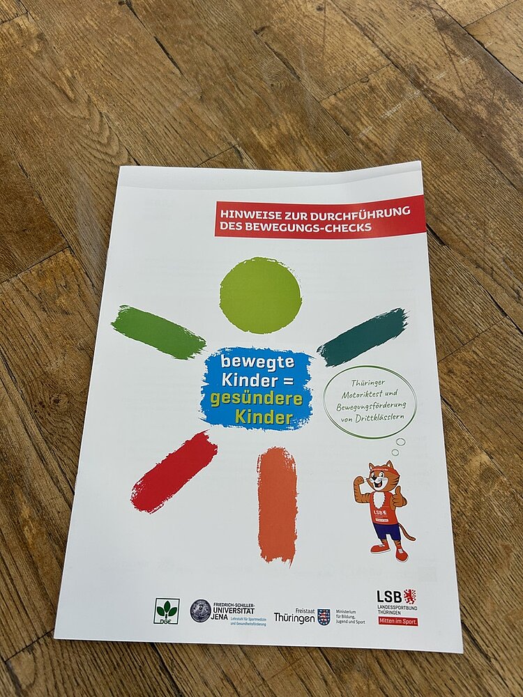 Informationsbroschüre des Thüringer Bewegungsprogramms Bewegte Kinder – Gesündere Kinder liegt auf einem hölzernen Hallenboden. Auf dem Titel sind farbige, pinselstrichartige Formen in Grün, Blau, Rot und Orange als stilisierte Figur angeordnet. Darüber steht der Hinweis Hinweise zur Durchführung des Bewegungs-Checks. Unten sind die Logos der Deutschen Gesellschaft für Ernährung, der Friedrich-Schiller-Universität Jena, des Freistaats Thüringen, des Thüringer Bildungsministeriums sowie des Landessportbunds Thüringen sichtbar. Die Broschüre ist Teil des verpflichtenden Bewegungsprogramms für alle dritten Klassen in Thüringen, das durch Auszubildende zu Erzieherinnen und Erziehern an der Euro Akademie Pößneck unterstützt wurde.