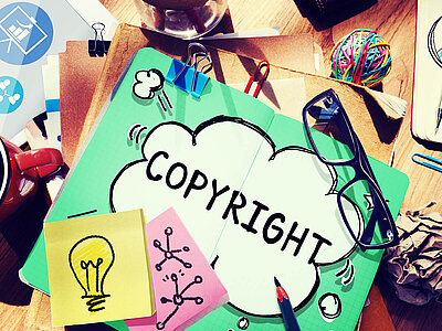 Ein grüner Ordner mit dem Wort 'COPYRIGHT' in einer Sprechblase liegt auf einem Schreibtisch. Auf dem Ordner befinden sich eine Brille, Büroklammern und ein gelber Notizzettel mit einer Glühbirne darauf. Um den Ordner herum liegen weitere Notizzettel, ein roter Tacker, ein Knäuel bunter Gummibänder und andere Büromaterialien.