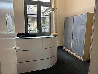 Ein leerer Empfangsbereich mit einem halbkreisförmigen Empfangstresen. Der Raum hat große Fenster, durch die man einen Blick auf ein Gebäude und Bäume im Freien erhaschen kann.