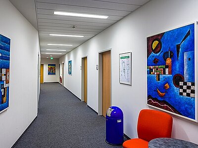 Blick in einen langen, hellen Flur der Euro Akademie Dresden. Links und rechts hängen großformatige und abstrakte, bunte Bilder.
