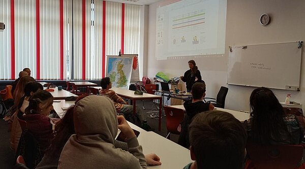 Career Day an der Euro Akademie In einem abgedunkelten Klassenzimmer lauschen Schülerinnen und Schüler einer Frau, die einen Vortrag hält. An der Wand sieht man die Präsentation, die den Vortrag unterlegt.