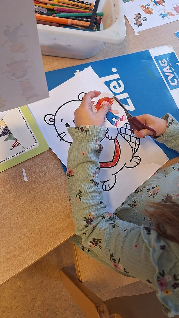 Nahaufnahme: Kind schneidet eine Papierfigur aus und arbeitet an einem Arbeitsblatt mit Tiermotiv; Stiftebox im Hintergrund.