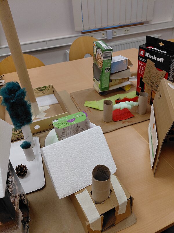 Mehrere aus Verpackungskartons und Papprollen gebaute Häuser und Türme stehen auf Kartonplatten auf einem Tisch; vorne ein kleines Haus mit weißem Styropordach und Papprollen-Schornstein, dahinter weitere fantasievolle Konstruktionen.