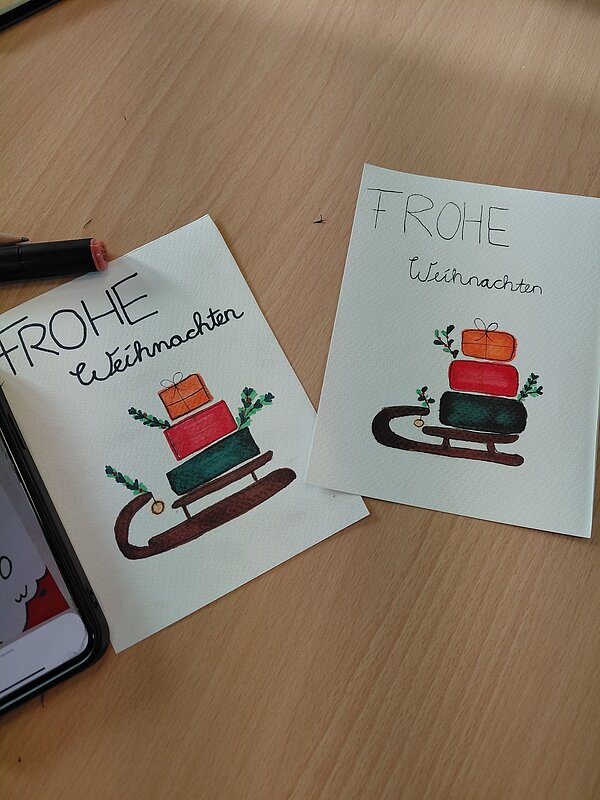 Zwei illustrierte Weihnachtskarten mit Geschenken auf einem Schlitten und der Aufschrift „FROHE Weihnachten“ liegen nebeneinander auf einem Holztisch.