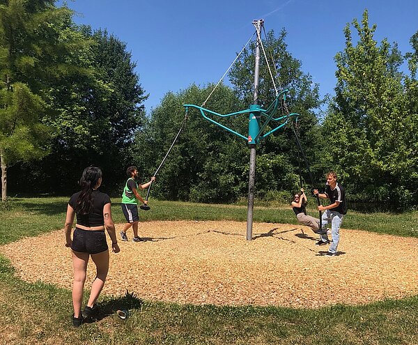  Drei junge Erwachsene befinden sich auf einer Schaukelkonstruktion auf einem Spielplatz, während eine Person im Vordergrund mit dem Rücken zur Kamera in Richtung der Schaukel läuft.