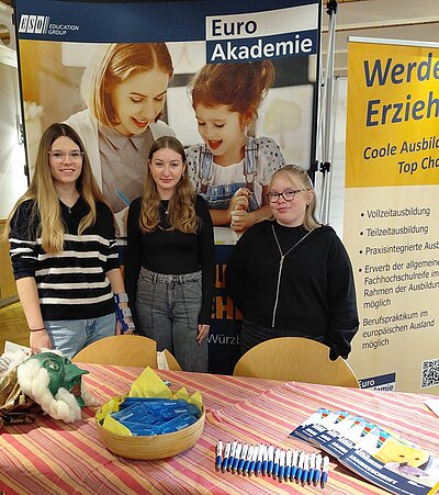 Drei weibliche Auszubildende stehen gut gelaunt am Messestand der Euro Akademie. Hinter ihnen ein großes Euro Akademie Roll-Up, vor ihnen ein auf dem Tisch liegen Kugelschreiber zum Mitnehmen sowie Informationsbroschüren.