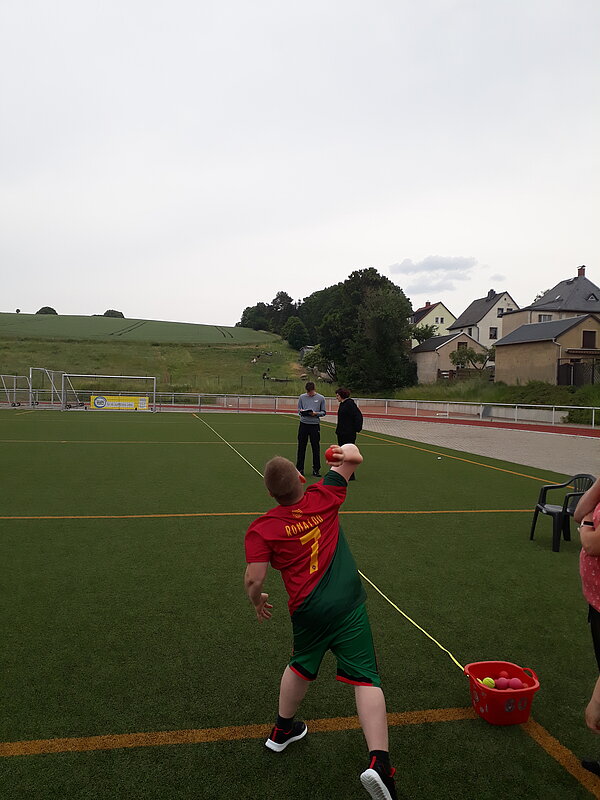 Unser Motto zum Sportfest: "Auf zu neuen Galaxien!" Ein Junge im roten Trikot mit der Aufschrift „Ronaldo“ wirft einen Ball auf einem Sportplatz. Ein Messband liegt am Boden.