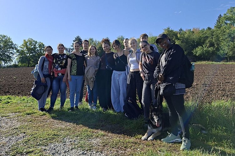 Zwölf Personen in lockerer Pose auf einem Feldweg, Arme teils umeinander gelegt, ein angeleinter Hund sitzt davor; im Hintergrund gepflügtes Ackerland und Bäume bei klarem Himmel.