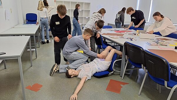 Schülerinnen und Schüler in einem großen Unterrichtsraum der Euro Akademie Hohenstein-Ernstthal, der mit Tischen, Stühlen und einem Regal ausgestattet ist, beschäftigen sich mit Lehrmaterial, das auf dem großen Tisch ausgebreitet ist. Drei Schüler beschäftigen sich mit einer lebensgroßen Dummy-Puppe, die auf einem umgekippten Stuhl platziert ist, und bringen ihn in die stabile Seitenlage. 