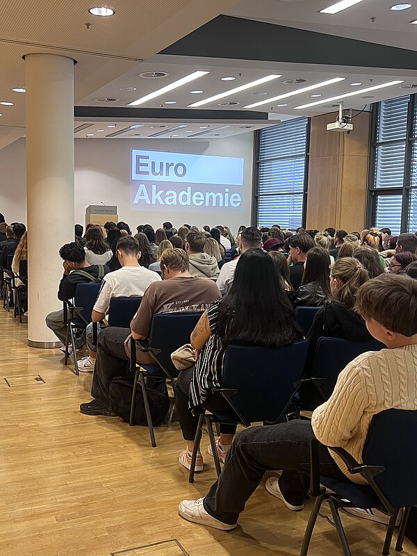 Zahlreiche Schüler*innen und Gäste sitzen in einem großen Veranstaltungssaal; auf der Leinwand ist das Logo der Euro Akademie zu sehen.