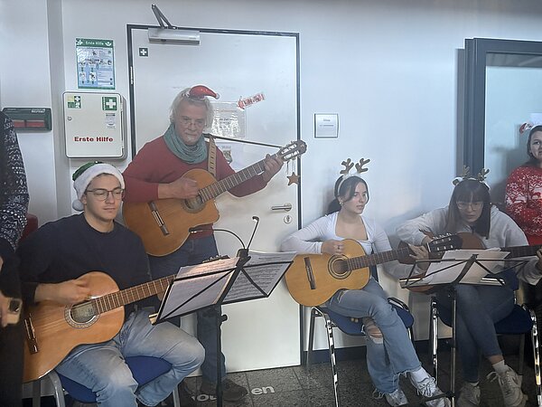 Mehrere Personen sitzen auf Stühlen und spielen Gitarren, einige tragen weihnachtliche Kopfbedeckungen, Notenständer stehen vor ihnen.