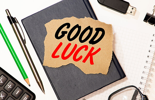 Ein Notizbuch mit einem aufgeklebten Zettel, auf dem in großen schwarzen und roten Buchstaben 'GOOD LUCK' steht, umgeben von Schreibutensilien wie Stiften, einem Taschenrechner, einer Brille und einem USB-Stick auf einem Schreibtisch.
