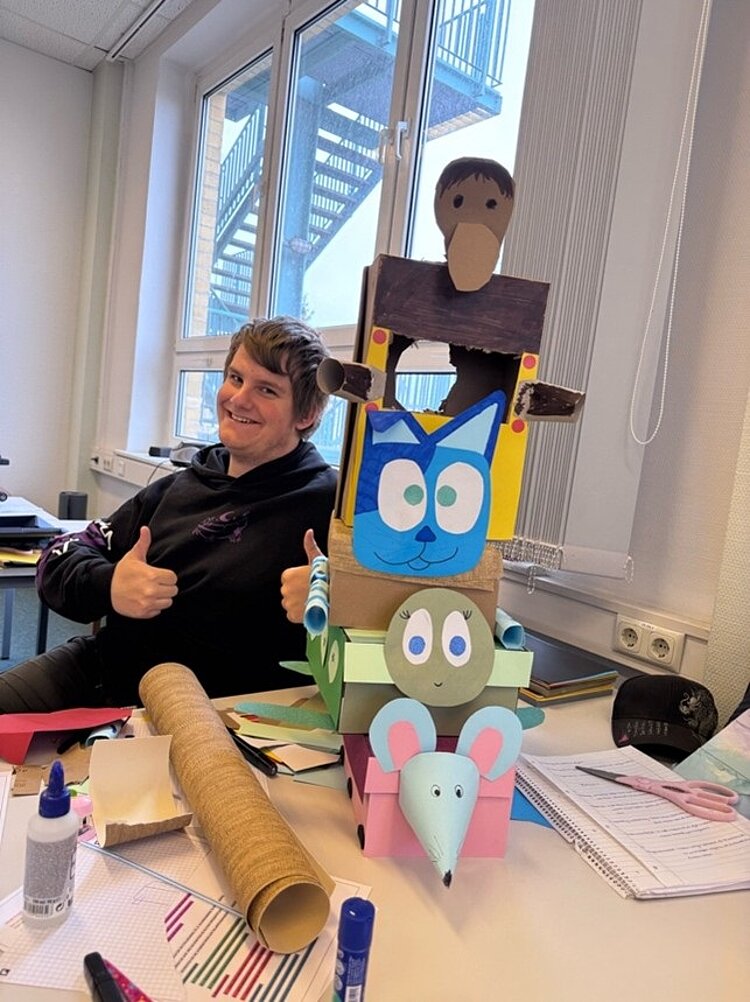 Auf einem Schreibtisch steht ein bunter Turm aus bemalten Kartons, die wie Tiere und Figuren gestaltet sind. Die unterste Ebene zeigt einen hellblauen Kopf mit großen Ohren und einer spitzen Schnauze, darüber befindet sich ein grünes Element mit einem weiteren Tiergesicht. Die mittlere Ebene zeigt ein blaues Gesicht mit großen, runden Augen und Katzenohren. Ganz oben ist ein brauner Karton mit einem rechteckigen Ausschnitt. Im Hintergrund sitzt ein Azubi der Euro Akademie Bitterfeld-Wolfen an einem Tisch und zeigt mit beiden Händen den Daumen nach oben. Auf dem Tisch liegen Bastelmaterialien wie Papierrollen, Kleber und Stifte. Die Szene spielt in einem hellen Raum mit großen Fenstern und Blick auf eine Metallaußentreppe.