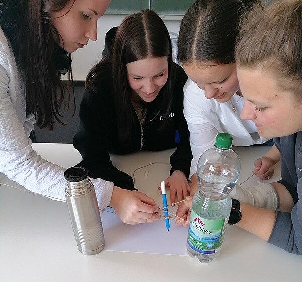 Teamwork Schülerinnen und Schüler sind über einen Tisch gebeugt und arbeiten an einer Gruppenaufgabe, in der sie etwas mithilfe einer Wasserflasche, Bändern und einem Stift bewerkstelligen müssen.
