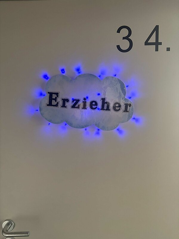 An einer hellen Tür ist die Nummer '34.' in großer schwarzer Schrift angebracht. Darunter befindet sich eine Wolkenform, die mit blauen LED-Lichtern umrandet ist. In der Wolke steht in schwarzer Schreibmaschinenschrift das Wort 'Erzieher'. An der linken unteren Ecke der Tür ist ein silberner Türgriff sichtbar. Das Bild entstand im Rahmen des Tages der offenen Tür an der Euro Akademie Zwickau.