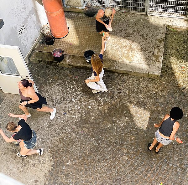 Blick von oben auf einen Innenhof mit nassem Kopfsteinpflaster, in dem mehrere Personen mit Wasserbomben spielen. Zwei Eimer mit bunten Wasserbomben stehen am Rand, einige Personen laufen lachend umher.