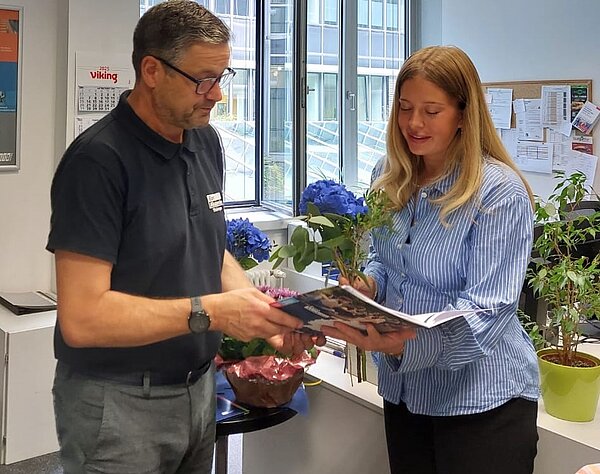 Der Mann überreicht einer jungen Frau im blauen Hemd eine Mappe und sie hält dabei einen Blumenstrauß.