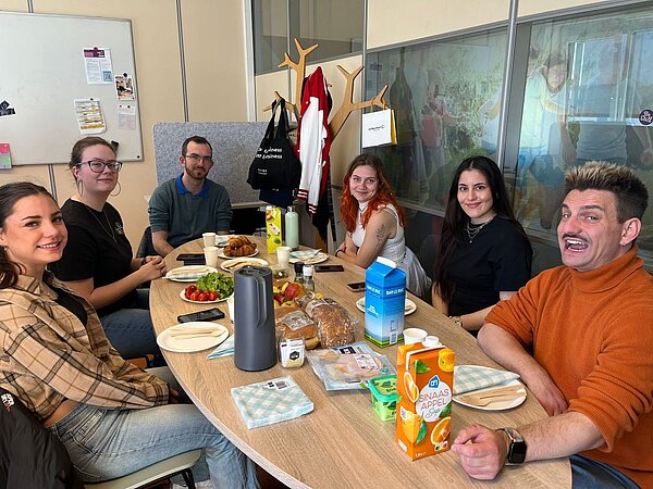 Sechs Personen sitzen fröhlich an einem runden Tisch und haben ein gemeinsames Frühstück oder eine Brotzeit. Auf dem Tisch stehen verschiedene Lebensmittel wie Brot, Obst, Saft, Milch und Aufstriche. Der Raum wirkt wie ein Gemeinschaftsraum.