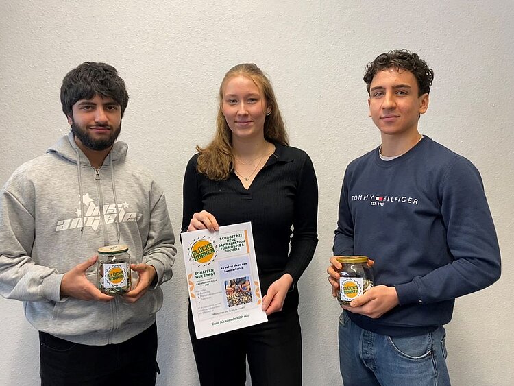 Sammelaktion: Kronkorken für den guten Zweck Drei junge Menschen stehen vor einer weißen Wand. Zwei von ihnen halten Gläser mit Kronkorken, die dritte Person hält ein Poster mit Informationen zur Sammelaktion „Glückskorken“. Alle schauen freundlich in die Kamera.