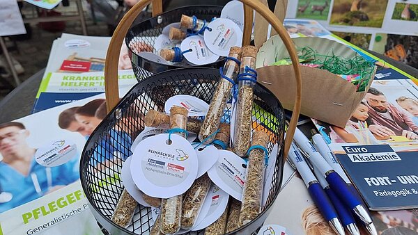 Ein Korb mit kleinen Glasflaschen, die Samen enthalten, steht auf einem Tisch neben Broschüren und Stiften. Die Flaschen tragen Etiketten mit der Aufschrift 'Klimaschule'.
