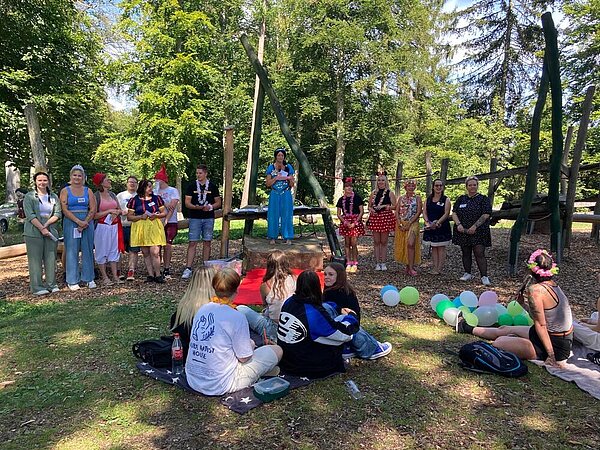 Eine Gruppe von jungen Erwachsenen in Disney-Kostümen sitzt und steht im Wald, während sie einer Frau im Kostüm zuhören, die etwas vorträgt.