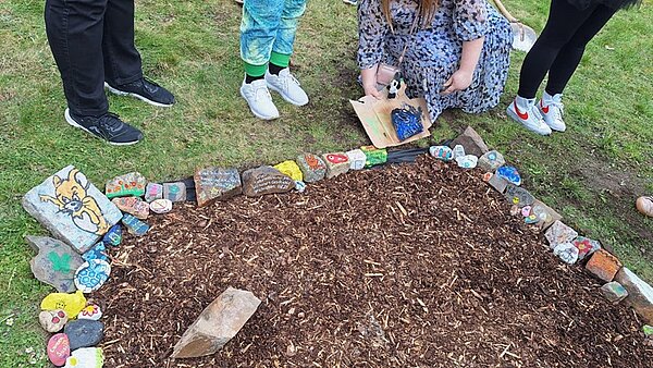 Mehrere Azubis stehen um ein mit Rindenmulch bedecktes Beet, das von bunt bemalten Steinen eingefasst ist. Die Steine zeigen verschiedene Motive, darunter ein Tiergesicht und abstrakte Muster. Eine Person hält eine bemalte Platte über das Beet. Der Rasen ist grün, und die Szene spielt im Freien.