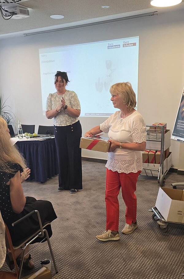 Eine Frau in einem weißen Oberteil und einer schwarzen Hose steht vor einem Projektor und klatscht in die Hände. Eine andere Frau in einem weißen Oberteil und roter Hose hält ein Paket mit rotem Band in den Händen. Im Hintergrund sind ein Tisch mit blauer Tischdecke, Stühle sowie ein Servierwagen mit weiteren Paketen zu sehen.