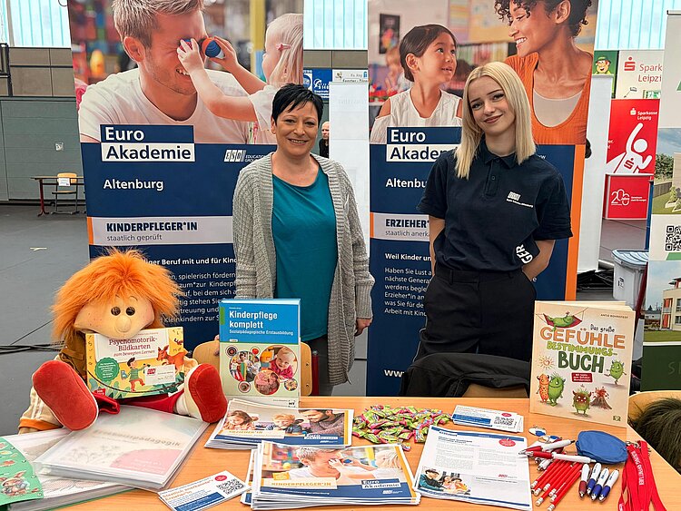 Zwei Personen stehen an einem Infostand der Euro Akademie Altenburg mit Roll-ups zu den Ausbildungen Kinderpflegerin und Erzieherin; auf dem Tisch liegen Infomaterialien, Stifte, Süßigkeiten, ein Buch zu Gefühlen und eine Stoffpuppe.