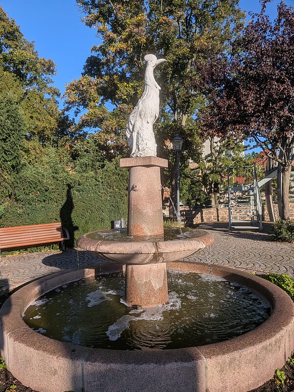 Steinbrunnen in einer grünen Parkanlage mit einer hohen Säule und einer weißen Vogelskulptur an der Spitze. Im Hintergrund stehen Bäume, eine Bank und eine Treppe.