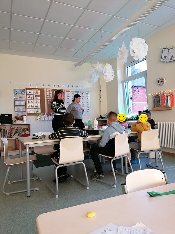 Engagiert und kreativ Zwei Auszubildende stehen am großen Basteltisch, an dem die Grundschüler sitzen und basteln.