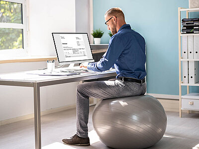 Auf dem Bild sitzt ein Mann auf einem Gymnastikball und arbeitet an einem Computer. Eine alternative Sitzoption für bessere Ergonomie.