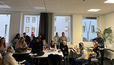 In einem Klassenzimmer sitzen auf den Tischen und um die Tische herum mehrere Schülerinnen und Schüler mit Textblättern in den Händen und singen. Ein Mann am Lehrerpult spielt dazu Gitarre.