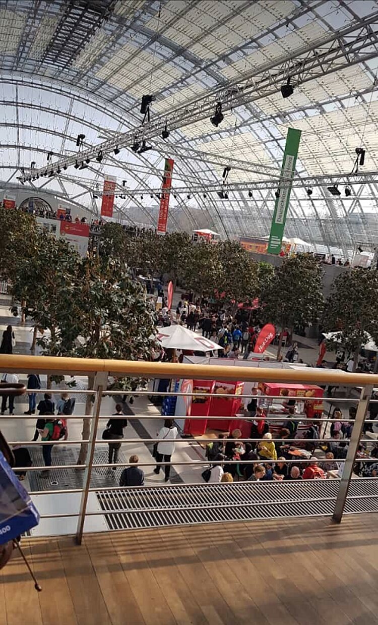 Zu Besuch auf der Buchmesse in Leipzig Blick von einem erhöhten Balkon in die Eingangshalle der Messe Leipzig, in der unter einer großen Glaskuppel Bäume wachsen.
